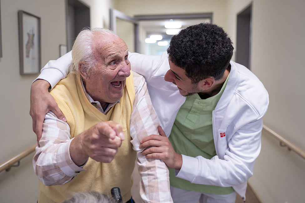 Pflegekraft umarmt lachenden Senior im Flur
