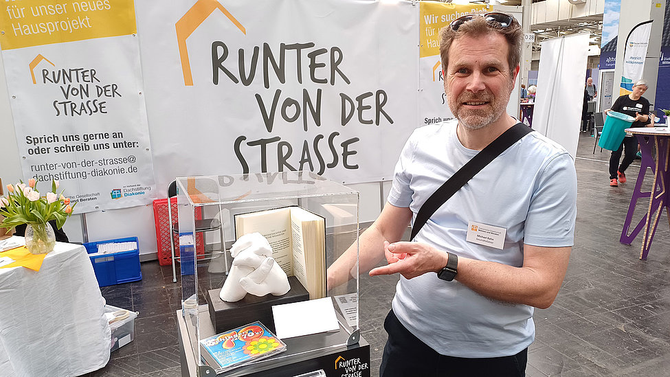 Person zeigt Exponat vor Messestand „Runter von der Straße“ mit Poster, Vitrine und Büchern.